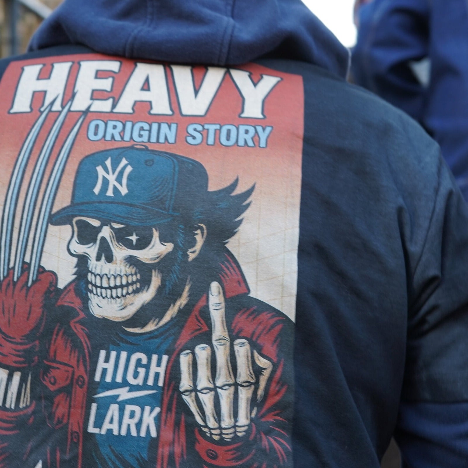 HEAVY x Highlark Wolverine Origin Story tee worn on stairs, back graphic in clear view — skeletal Wolverine in Yankees fitted, claws and middle finger raised, HIGH/LARK wordmark at bottom