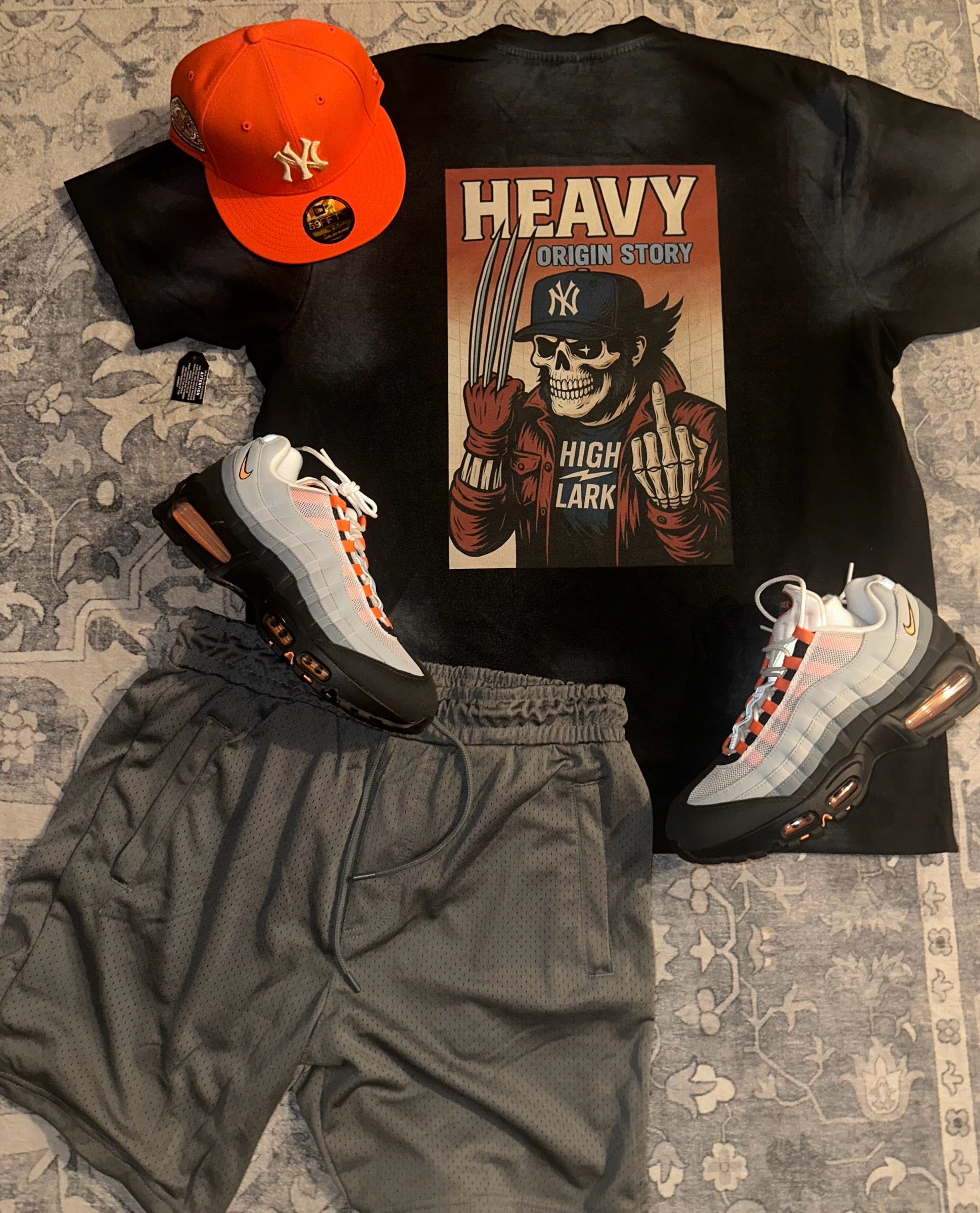 HEAVY x Highlark Wolverine Origin Story tee styled flat lay with orange Yankees fitted, Air Max 95 sneakers, and shorts on patterned rug.