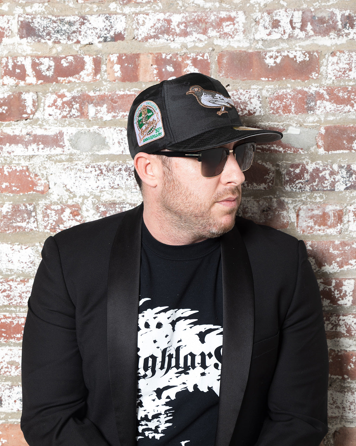 Highlark Baltimore Orioles 3.0 fitted cap on model against brick wall, 30th Anniversary sidepatch visible with Highlark Collective tee and uxedo blazer styling