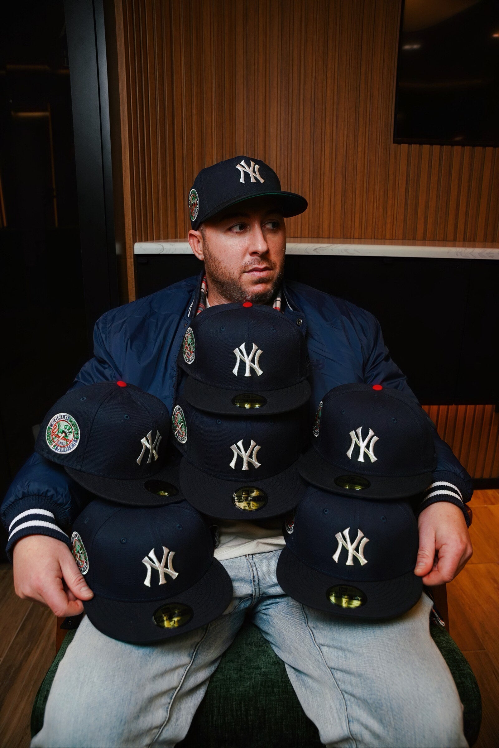 Mitch Highlark seated in restaurant booth wearing navy bomber and Timeless Yankees fitted, holding five additional Timeless Yankees fitteds stacked on his lap, against wood paneling
