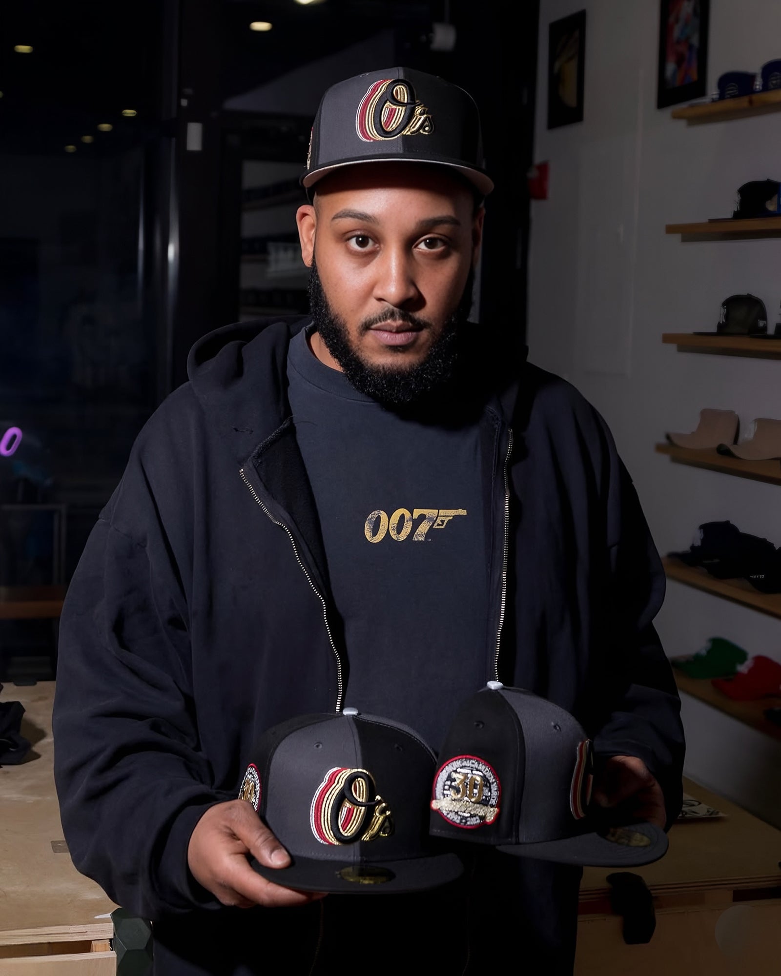 Tone holding the Highlark Baltimore Orioles 007 James Bond fitted hat and the Orioles 3.0 inside Home Game NY, wearing a black 007 logo hoodie